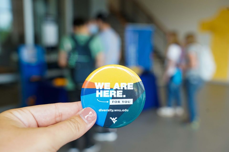 A hand holding up a promotional pin for the division of diversity, equity and inclusion that reads 'We are here for you.' in white text against a rainbow background.