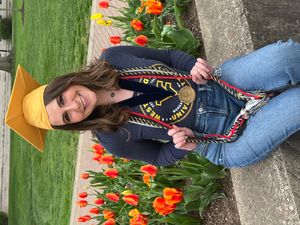 Lauren Volk sits in front of tulips wearing a gold mortarboard, medallion and three graduation cords